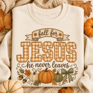 以下が含まれることがあります： 「fall for JESUS he never leaves」という刺繍が施されたクリーム色のクルーネックトレーナー。文字にはベージュのギンガムチェック柄があしらわれ、オレンジと緑のカボチャ、どんぐり、緑色のリボンの刺繍がデザインされています。