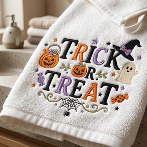 Peut inclure: Une serviette à main en tissu éponge blanc avec des broderies sur le thème d'Halloween, incluant le texte Trick or Treat, deux citrouilles d'Halloween, un chapeau de sorcière noir, un petit fantôme, une toile d'araignée et des bonbons colorés.