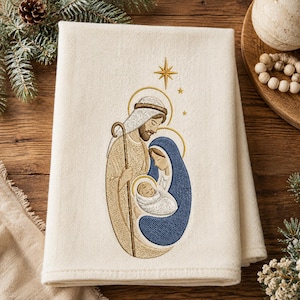 May include: A cream-coloured fabric towel featuring an embroidered nativity scene. The design depicts a figure in tan robes holding a staff, a figure in a blue head covering, and a swaddled infant, all beneath a gold embroidered star.