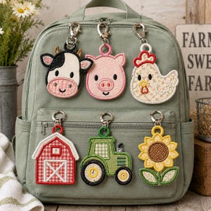 以下が含まれることがあります： セージグリーンのバックパックに、6つの刺繍入りキーホルダーがファスナーに取り付けられています。キーホルダーは牛、豚、鶏、赤い納屋、緑のトラクター、黄色いひまわりをモチーフにしており、それぞれ銀色の金属製ロブスタークラスプで固定されています。