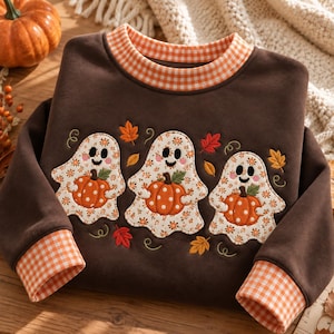 Könnte beinhalten: Ein dunkelbraunes Langarm-Sweatshirt mit einem orange-weiß karierten Kragen und Bündchen. Auf der Vorderseite befinden sich drei gestickte Geisterfiguren, die jeweils einen orangefarbenen, gepunkteten Kürbis halten, umgeben von kleinen Herbstblättern.