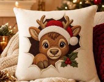 May include: A square cream-colored throw pillow featuring an embroidered design of a brown reindeer wearing a red and white Santa hat. The reindeer appears to be emerging from a dark hole, accented with green holly leaves and red berries.