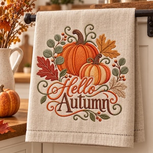 May include: A beige linen-look kitchen towel featuring an embroidered design of two orange pumpkins, autumn leaves, and berries. The text "Hello Autumn" is stitched in a decorative script font in the center of the pumpkin and foliage arrangement.