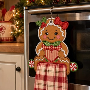 May include: A gingerbread person-shaped kitchen towel holder made of embroidered fabric. It features a red bow, holly leaves, and a red and white plaid heart. A red and white plaid towel hangs from the bottom, flanked by two peppermint candy accents.
