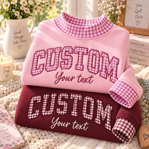 Puede incluir: Dos sudaderas de manga larga, una rosa claro y otra granate oscuro, con cuello alto de patrón de cuadros vichy rosa y blanco. La palabra CUSTOM está bordada en cuadros en el pecho, con el texto Your text escrito debajo.