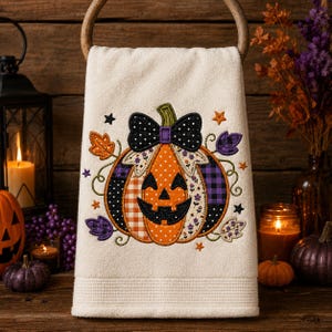 May include: A cream-colored hand towel featuring an embroidered jack-o'-lantern pumpkin with a black polka-dot bow. The pumpkin design incorporates orange, purple, and black fabric patterns, surrounded by embroidered autumn leaves and small stars.