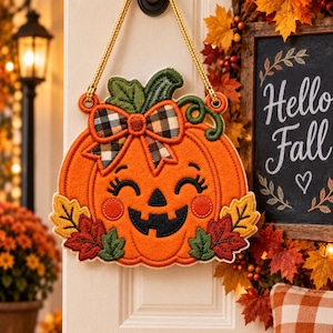 Può includere: Una decorazione da parete in feltro a forma di zucca con un volto sorridente, occhi chiusi, guance rosate e naso e bocca neri. La zucca è decorata con un fiocco a quadri bianco e nero, foglie verdi e fogliame autunnale rosso e giallo.