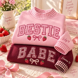 May include: Two long-sleeve sweatshirts, one light pink and one dark maroon, featuring a high collar with a pink and white gingham pattern. The light pink sweatshirt displays the text BESTIE, while the maroon sweatshirt displays the text BABE.
