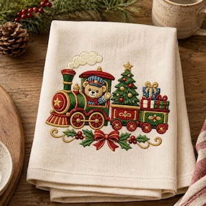 May include: A cream-coloured kitchen towel featuring an embroidered design of a green and red steam train driven by a teddy bear. The train carries a decorated Christmas tree and stacked gift boxes, accented by a red bow and holly leaves.
