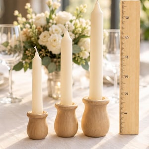 Puede incluir: Tres velas cónicas de color blanco roto en candelabros de madera clara de diferentes alturas. Una regla de madera está colocada junto a la vela más alta, mostrando una altura de aproximadamente 16,5 cm para el conjunto más alto.