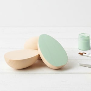 Peut inclure: Des formes d'œufs en bois, dont une peinte en vert clair, sont disposées sur une surface blanche. Des pots de peinture verte et lilas, un pinceau, une spatule et une éponge suggèrent un projet de bricolage ou d'artisanat.