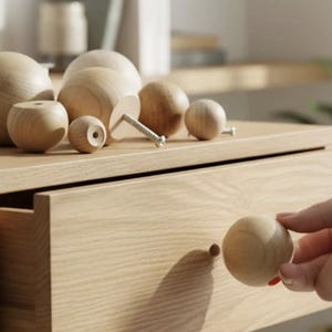 Puede incluir: Una colección de pomos esféricos de madera de color claro en varios tamaños. Varios pomos presentan una parte posterior plana con un orificio central, mientras que uno incluye un tornillo de metal. Un pomo esférico se sostiene cerca de un cajón.