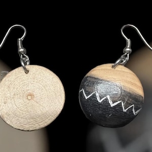 May include: A pair of drop earrings featuring circular wooden beads suspended from silver-toned metal hooks. One bead displays a natural light wood grain, while the other is partially painted black with a white zigzag line across the center.