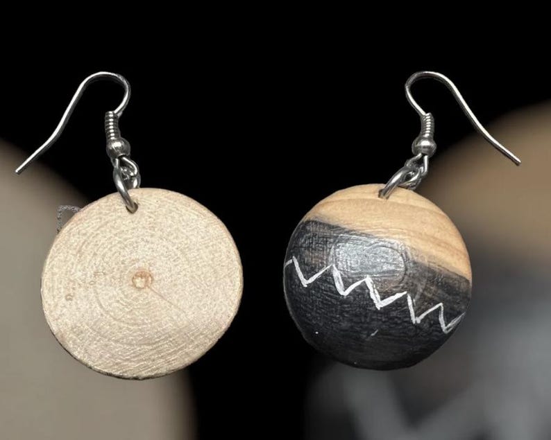 May include: A pair of drop earrings featuring circular wooden beads suspended from silver-toned metal hooks. One bead displays a natural light wood grain, while the other is partially painted black with a white zigzag line across the center.