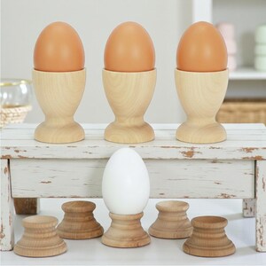 May include: Three light brown wooden egg cups holding brown eggs are arranged on a white wooden riser. Below the riser, a single white egg sits in a matching wooden cup, surrounded by four empty wooden egg cups on a white surface.
