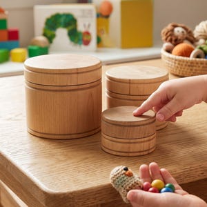 Peut inclure: Trois boîtes cylindriques en bois avec couvercles, disposées par taille décroissante. Les boîtes présentent une finition en bois clair lisse avec des détails rainurés horizontaux autour des côtés de chaque couvercle et de la base.