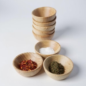Könnte beinhalten: Sechs kleine, hellfarbene Holzschalen. Drei Schalen sind mit Gewürzen gefüllt: rote Chiliflocken, weißes Salz und grüne Kräuter. Die Schalen sind auf einer weißen Oberfläche angeordnet, ideal zum Servieren von Gewürzen.