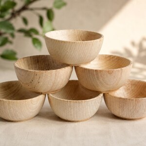 May include: A stack of six small, light-colored wooden bowls with a visible natural grain pattern. The bowls are arranged in a pyramid formation, featuring a smooth, rounded interior and a simple, circular base for each individual piece.