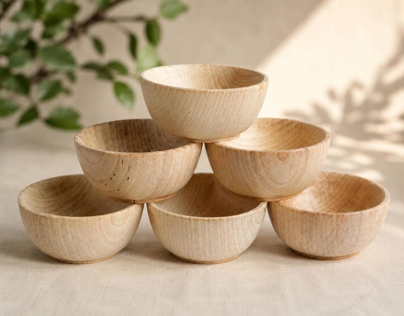 May include: A stack of six small, light-colored wooden bowls with a visible natural grain pattern. The bowls are arranged in a pyramid formation, featuring a smooth, rounded interior and a simple, circular base for each individual piece.