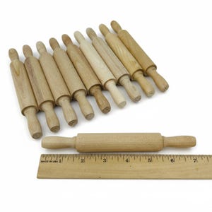 May include: A collection of wooden rolling pins in various shades of light brown, arranged on a white surface. One rolling pin is positioned horizontally next to a wooden ruler, marked with inches. The rolling pins are of different sizes and shapes.