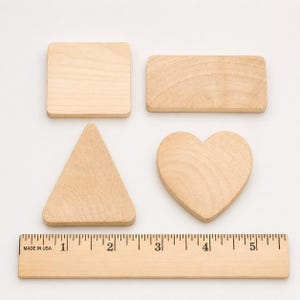 May include: Four light-colored wooden shapes including a square, a rectangle, a triangle, and a heart, arranged above a wooden ruler that measures up to 5 inches, showing the square is approximately 2 inches wide and the heart is 2 inches wide.