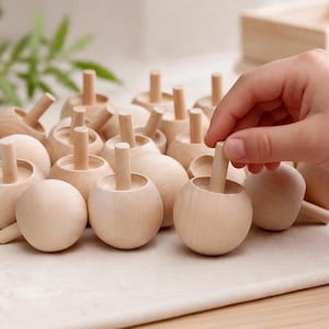 May include: A collection of light-colored wooden spinning tops, each featuring a smooth, rounded base and a central vertical handle. The tops are arranged in a group, with one being held by a hand to demonstrate the functional design.