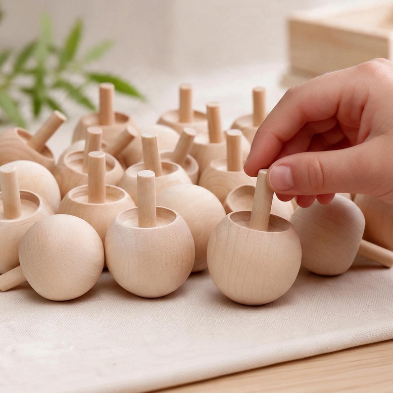 May include: A collection of light-colored wooden spinning tops, each featuring a smooth, rounded base and a central vertical handle. The tops are arranged in a group, with one being held by a hand to demonstrate the functional design.