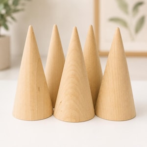 May include: A set of five smooth, light-coloured wooden cones arranged in a cluster. Each cone features a natural wood grain pattern with a wide circular base that tapers to a point at the top. The objects are solid, unpainted, and have a matte finish.