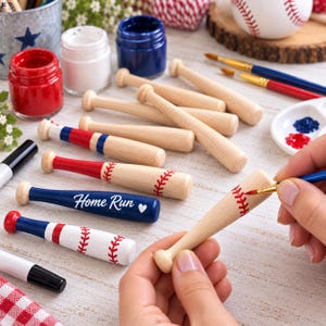 May include: A collection of small wooden baseball bats, some plain and some painted with red and blue stripes or red baseball stitching patterns. One blue bat features the white text Home Run and a heart symbol. Paint jars and brushes are present.