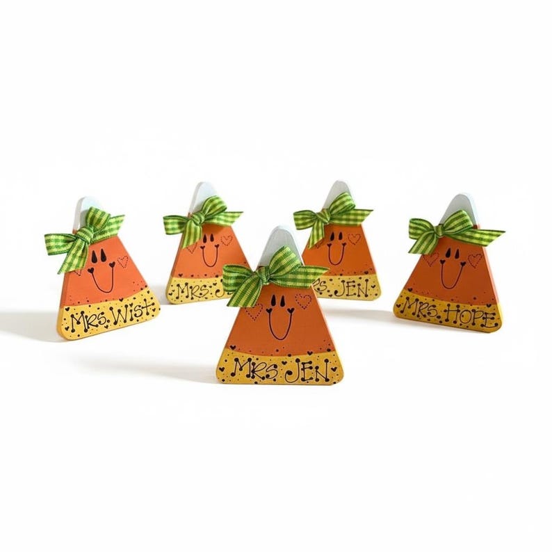 May include: Five triangular wooden candy corn decorations in orange and yellow with hand-painted faces and green gingham bows. Each piece features a name written in black script: Mrs. Wist, Mrs. J, Mrs. Jen, and Mrs. Hope, with heart accents.