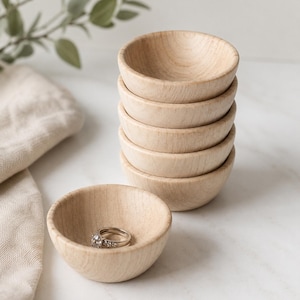 May include: A stack of five small, round, light-colored wooden bowls with a smooth finish. A single silver ring with small clear stones is placed inside an additional matching wooden bowl positioned in the foreground of the stack.