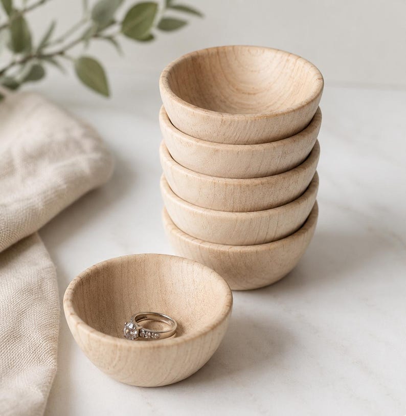 May include: A stack of five small, round, light-colored wooden bowls with a smooth finish. A single silver ring with small clear stones is placed inside an additional matching wooden bowl positioned in the foreground of the stack.