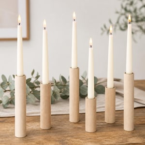 May include: Six tall, slender, off-white taper candles are each set into individual, cylindrical, light-toned wooden candle holders of varying heights. Each candle is lit, producing a small, warm flame at the top of the wax taper.
