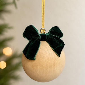 May include: A spherical light-colored wooden ornament featuring a dark green velvet bow attached to the top. A thin gold ribbon loop is secured to a silver metal eyelet at the center of the bow for hanging the round wooden decoration.