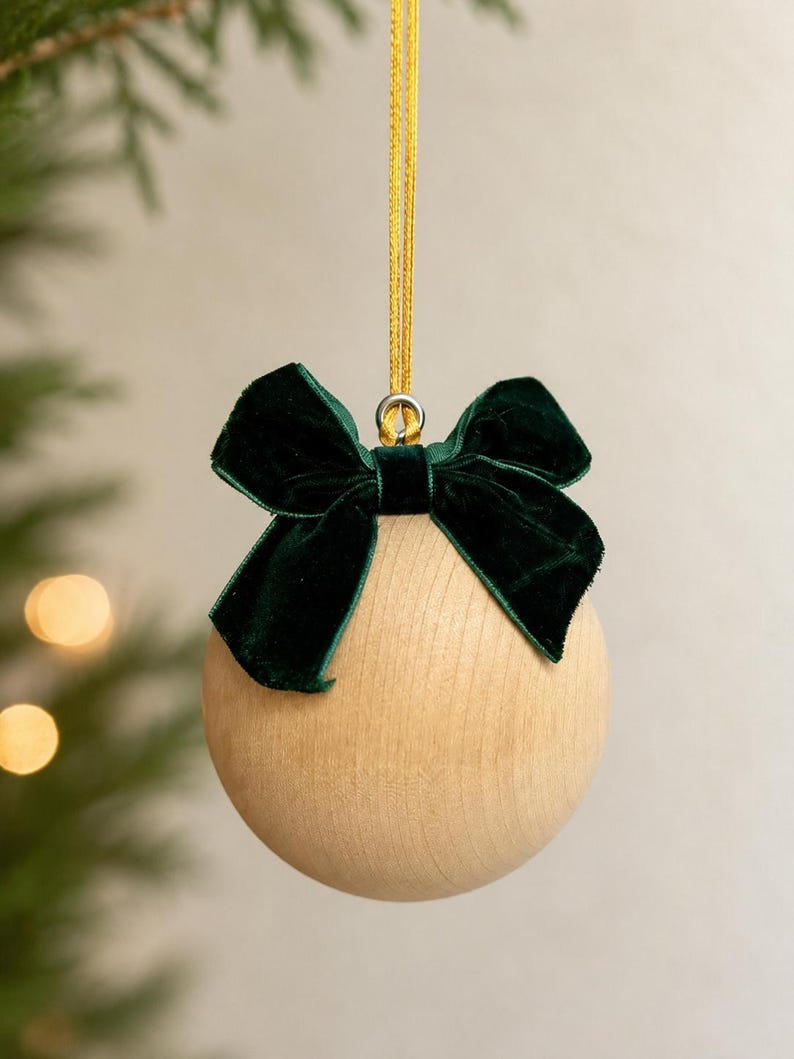 May include: A spherical light-colored wooden ornament featuring a dark green velvet bow attached to the top. A thin gold ribbon loop is secured to a silver metal eyelet at the center of the bow for hanging the round wooden decoration.
