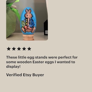 May include: A light blue wooden egg painted with a brown rabbit wearing a blue jacket and holding a green vegetable. The egg is positioned on a small, natural-colored wooden stand. The rabbit has dark outlines and is depicted in a seated position.