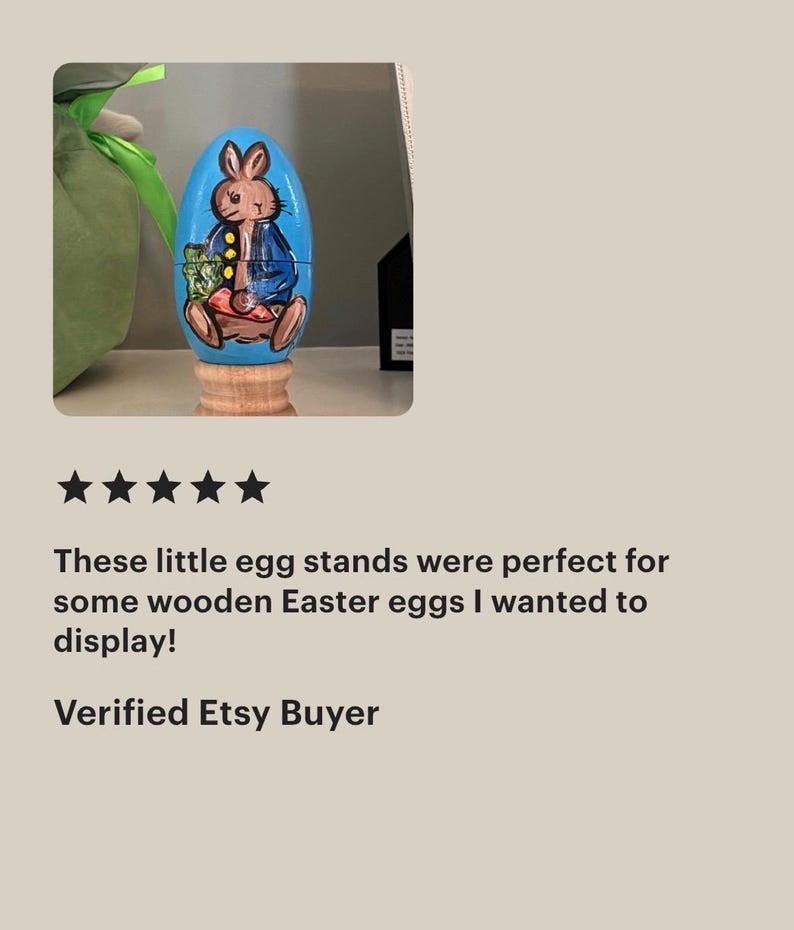 May include: A light blue wooden egg painted with a brown rabbit wearing a blue jacket and holding a green vegetable. The egg is positioned on a small, natural-colored wooden stand. The rabbit has dark outlines and is depicted in a seated position.