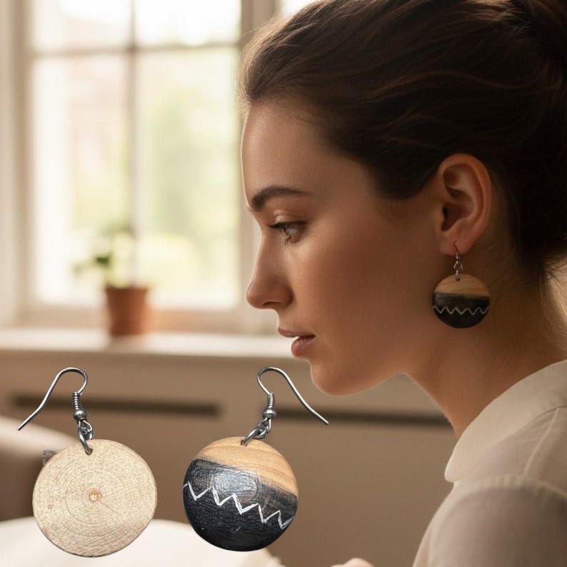 May include: Round wooden drop earrings featuring a natural light wood grain on the top half and a dark charcoal bottom half with a white zigzag line pattern. The earrings hang from silver-toned metal hooks and display a circular wood slice design.