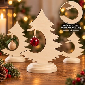 May include: A light-colored wooden Christmas tree cutout standing on a circular base. The tree features a large circular opening in the center for hanging ornaments. A shiny red jingle bell hangs from the top of the opening by a twine string.