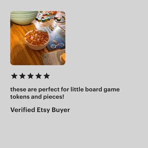 May include: A small, round, light-colored wooden bowl filled with numerous translucent, amber-colored square tokens. The bowl sits on a wooden surface next to a board game featuring various colorful circular markers and game cards.