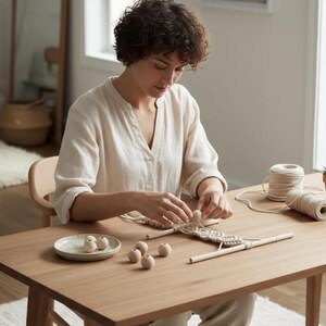 May include: A macrame wall hanging project in progress featuring natural cotton cord, light-colored wooden dowels, and several smooth, round wooden beads. The project is laid out on a wooden table with a small ceramic dish holding extra beads.