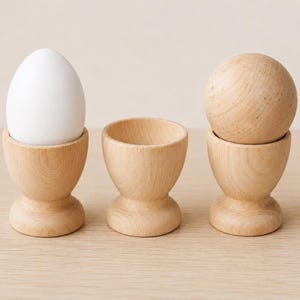 May include: Three light-colored wooden egg cups arranged in a row. The left cup holds a smooth white egg, the middle cup is empty, and the right cup holds a round, light-colored wooden sphere that matches the material of the cups.