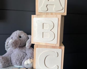 May include: Three stacked light-colored wooden blocks featuring large, raised white letters A, B, and C. Beside them sits a gray plush elephant toy and four smaller wooden blocks arranged in a row, spelling out the name JACK in colorful letters.