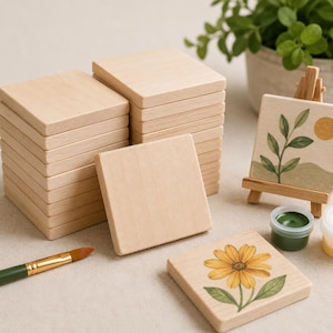 May include: A stack of square light-colored wooden tiles, one single square wooden tile, and one square wooden tile featuring a yellow flower illustration. A small wooden easel holds a tile with a leaf and sun design, next to a green paint pot and brush.