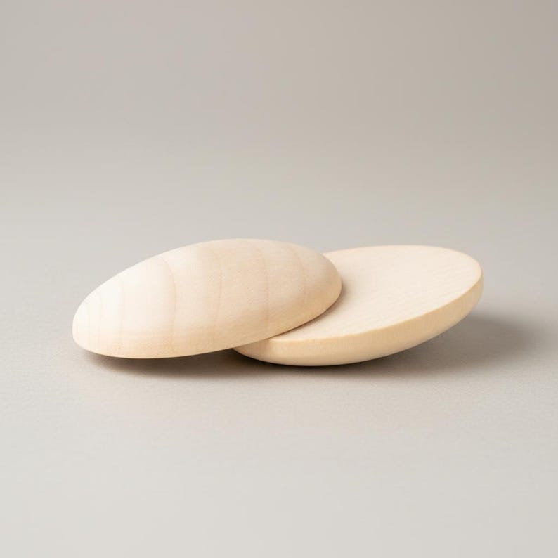 May include: Two smooth, unfinished light-colored wooden discs with a visible natural grain pattern. The circular wooden pieces are positioned overlapping each other, showcasing a rounded, convex shape on the top surface and a flat base.