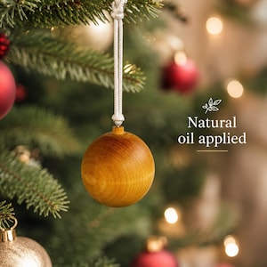 May include: A spherical wooden Christmas ornament with a smooth, light brown finish and a natural oil applied. The ornament hangs from a white braided cord and features the text Natural oil applied printed in white on the right side.