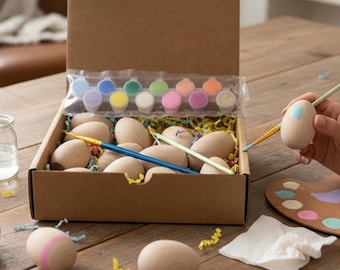 May include: A craft kit for decorating wooden Easter eggs. The kit includes a cardboard box filled with unfinished wooden eggs, paint pots in various colors, and paintbrushes. One egg is being painted with a light blue color. A wooden palette and a white paper towel are also visible.