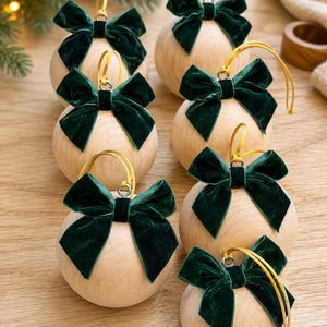 May include: A set of six spherical wooden ornaments, each featuring a dark green velvet bow attached to a gold hanging cord. The natural light-colored wood grain is visible on the smooth, round surface of every individual ornament.