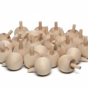 May include: A collection of numerous unfinished light-colored wooden spinning tops. Each toy features a smooth, rounded, spherical base with a straight, cylindrical handle protruding from the center, showcasing the natural grain of the wood.