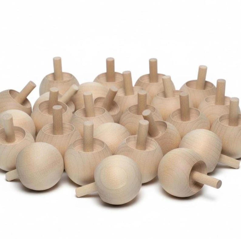 May include: A collection of numerous unfinished light-colored wooden spinning tops. Each toy features a smooth, rounded, spherical base with a straight, cylindrical handle protruding from the center, showcasing the natural grain of the wood.